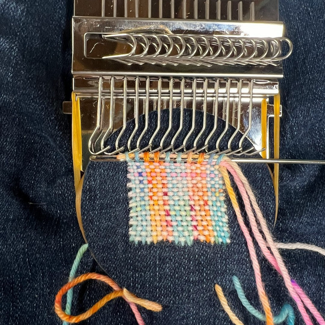 A darning loom clamped over dark denim with a finished woven patch in pastel and orange yarn still on the hooks, tapestry needle at the last row