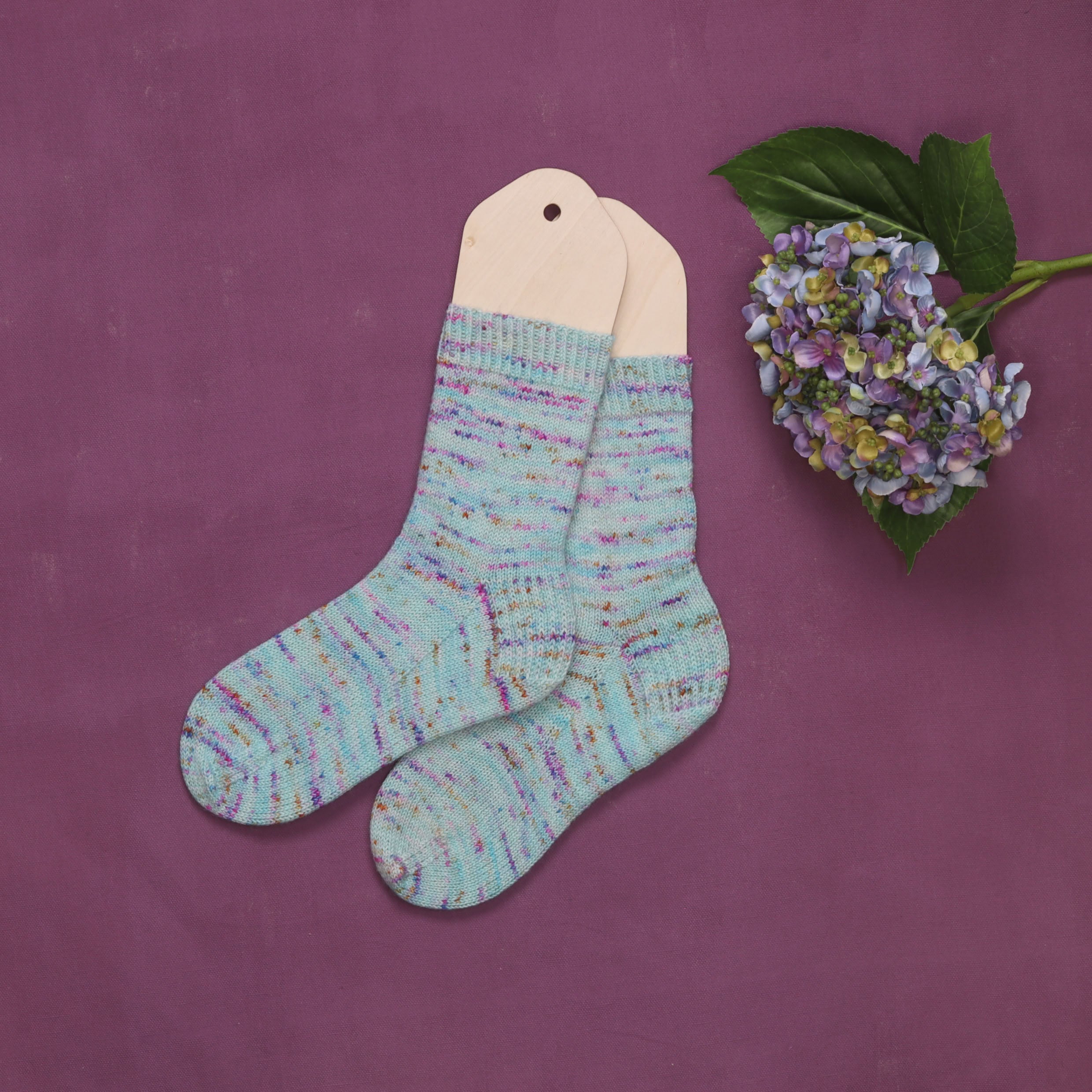 A finished pair of pale aqua speckled hand-knitted socks on wooden sock blockers, with a hydrangea sprig