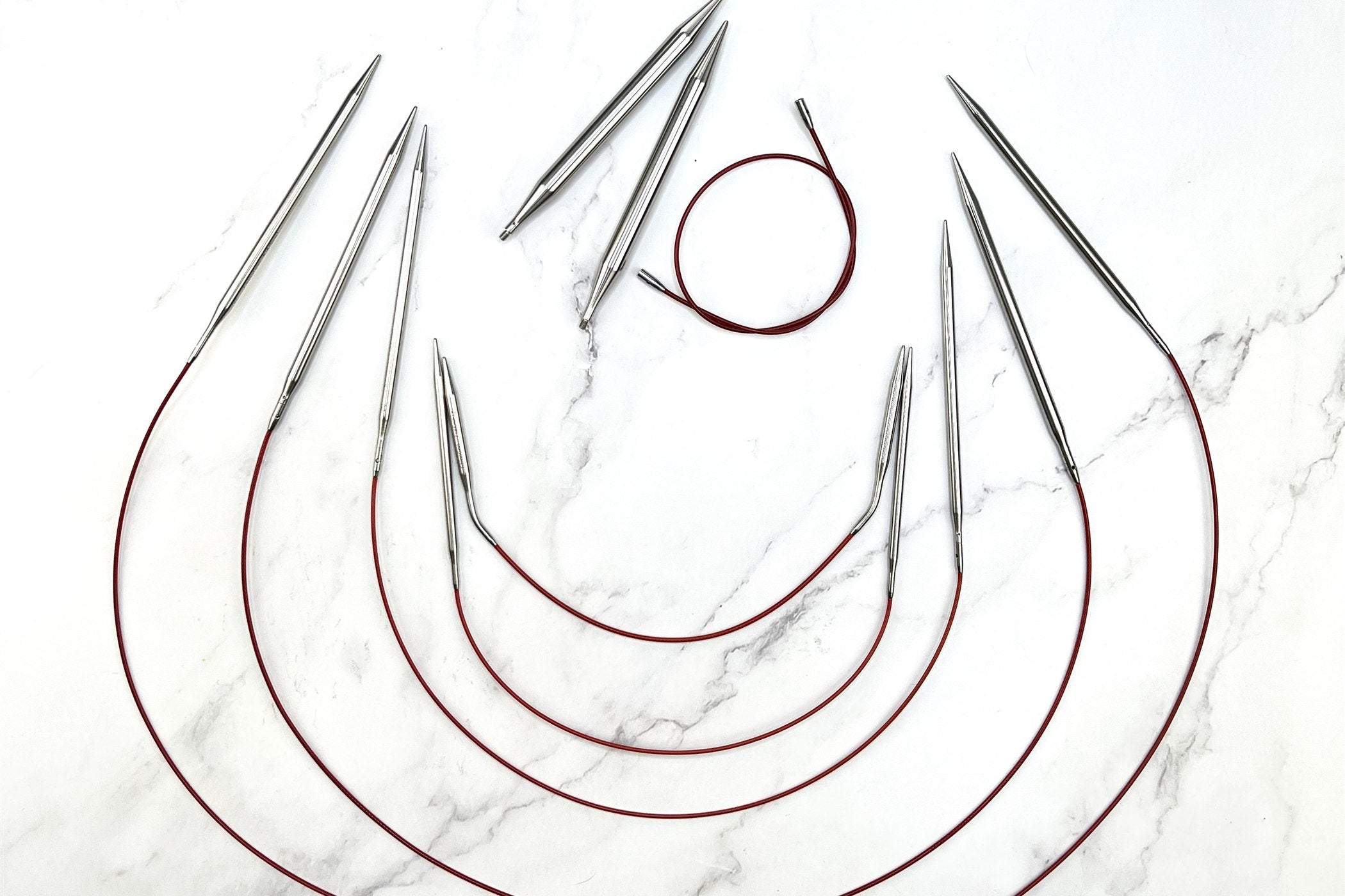 Five ChiaoGoo circular knitting needles in graduated cable lengths fanned out on white marble
