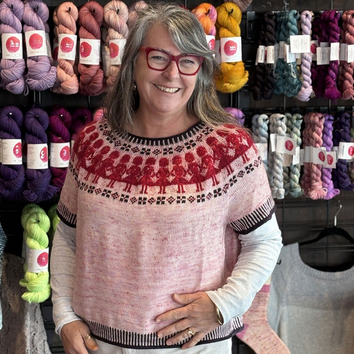 Woman wearing a pink sweater with red patterns in front of yarn shelves.
