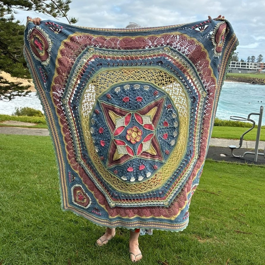 Universe in Bloom Blanket crochet kit – finished blanket in Gum Blossom held up by the sea