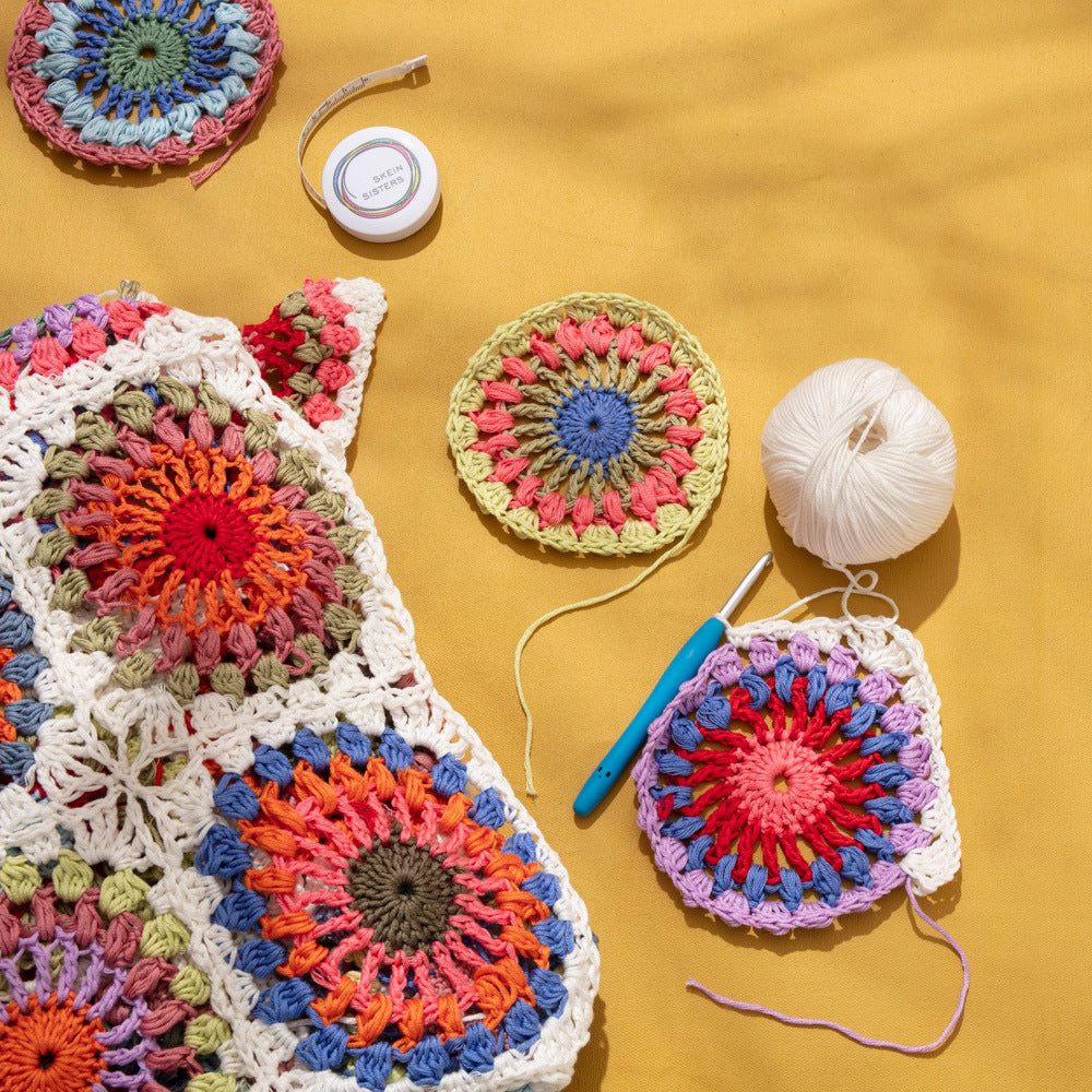 Colourful crocheted mandala granny squares in progress on a mustard cloth, with a ball of cream cotton, a teal crochet hook and a Skein Sisters tape measure