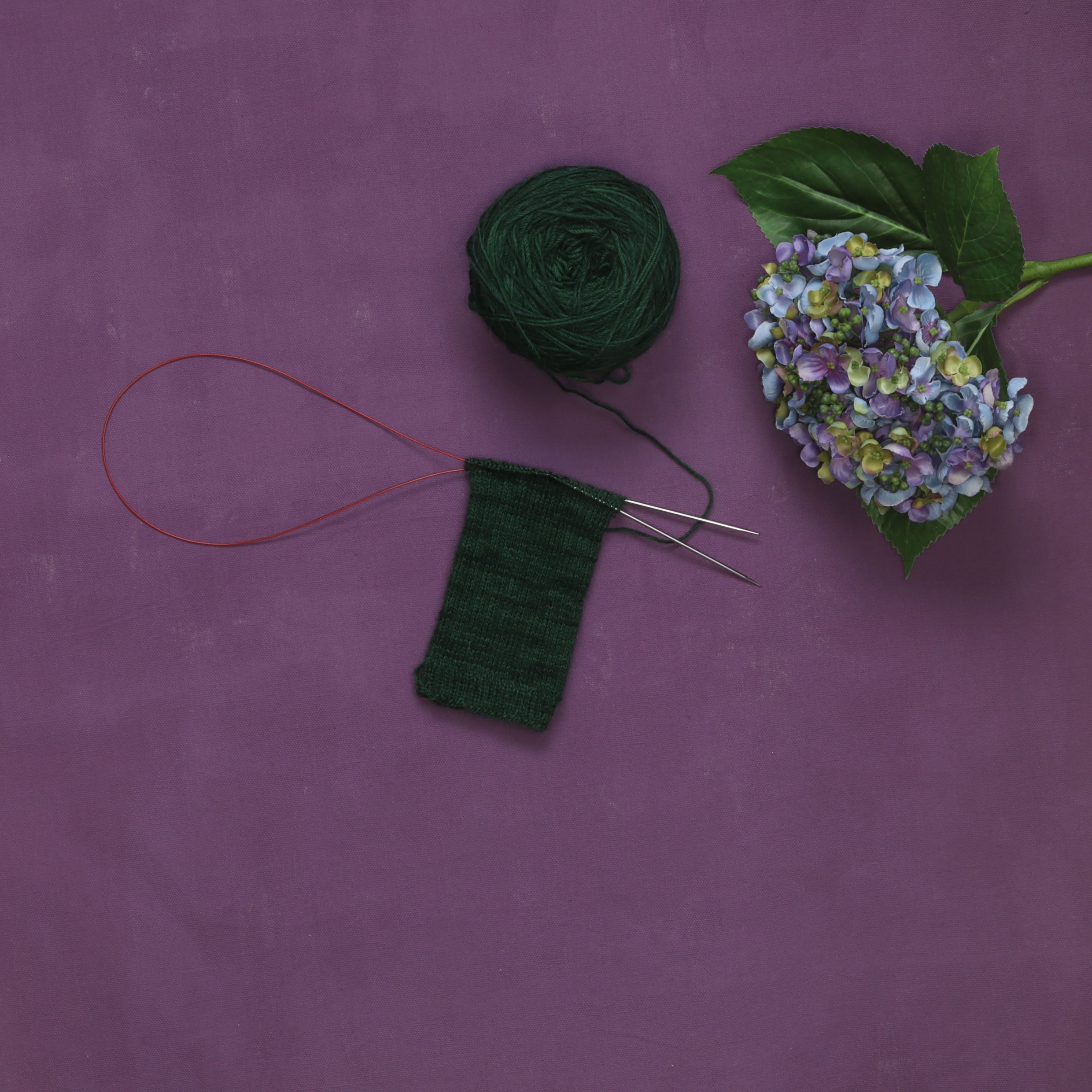 A dark green sock being knitted from the top down on a circular needle, with a cake of the same yarn