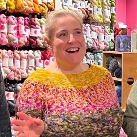 Person wearing a colorful knitted sweater in front of yarn shelves