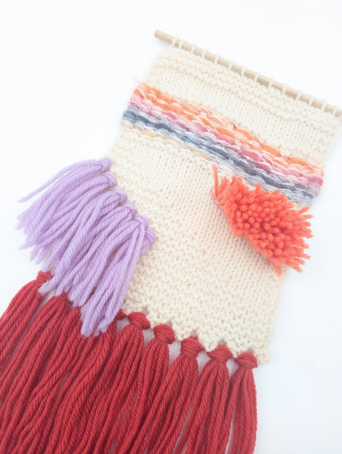 Knitted shaggy wall hanging on a dowel with lilac and red long fringe and an orange shaggy tuft